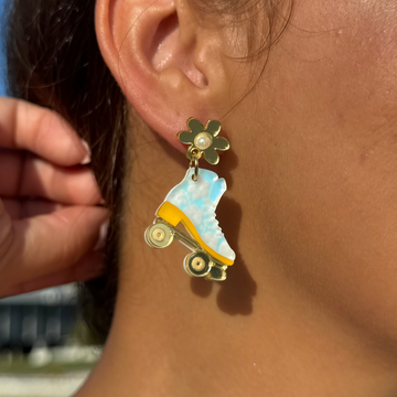 Handmade blue and yellow acrylic roller skate earring with gold daisy stud, worn on ear in sunlight.