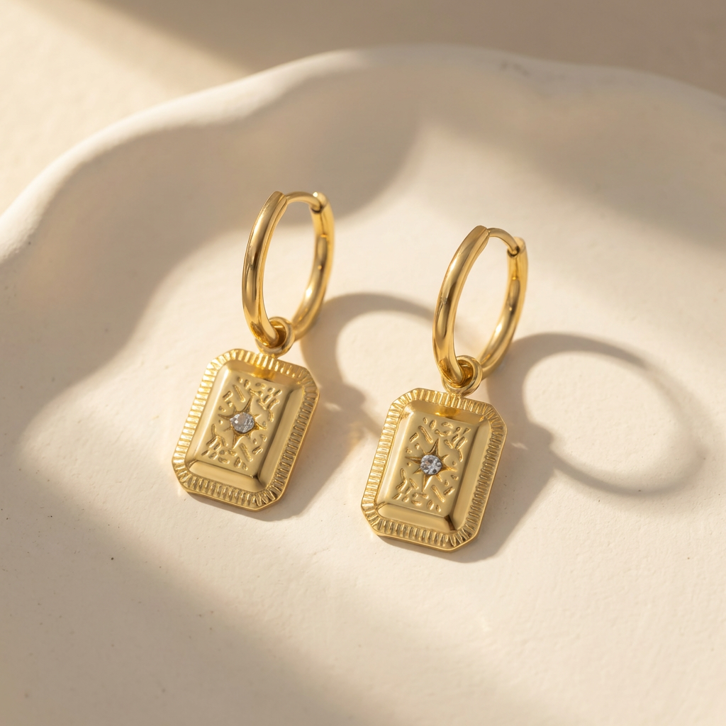 Gold tone stainless steel stamped charm huggie hoop earrings flat lay on sculptural ceramic edge backdrop with soft shadows
