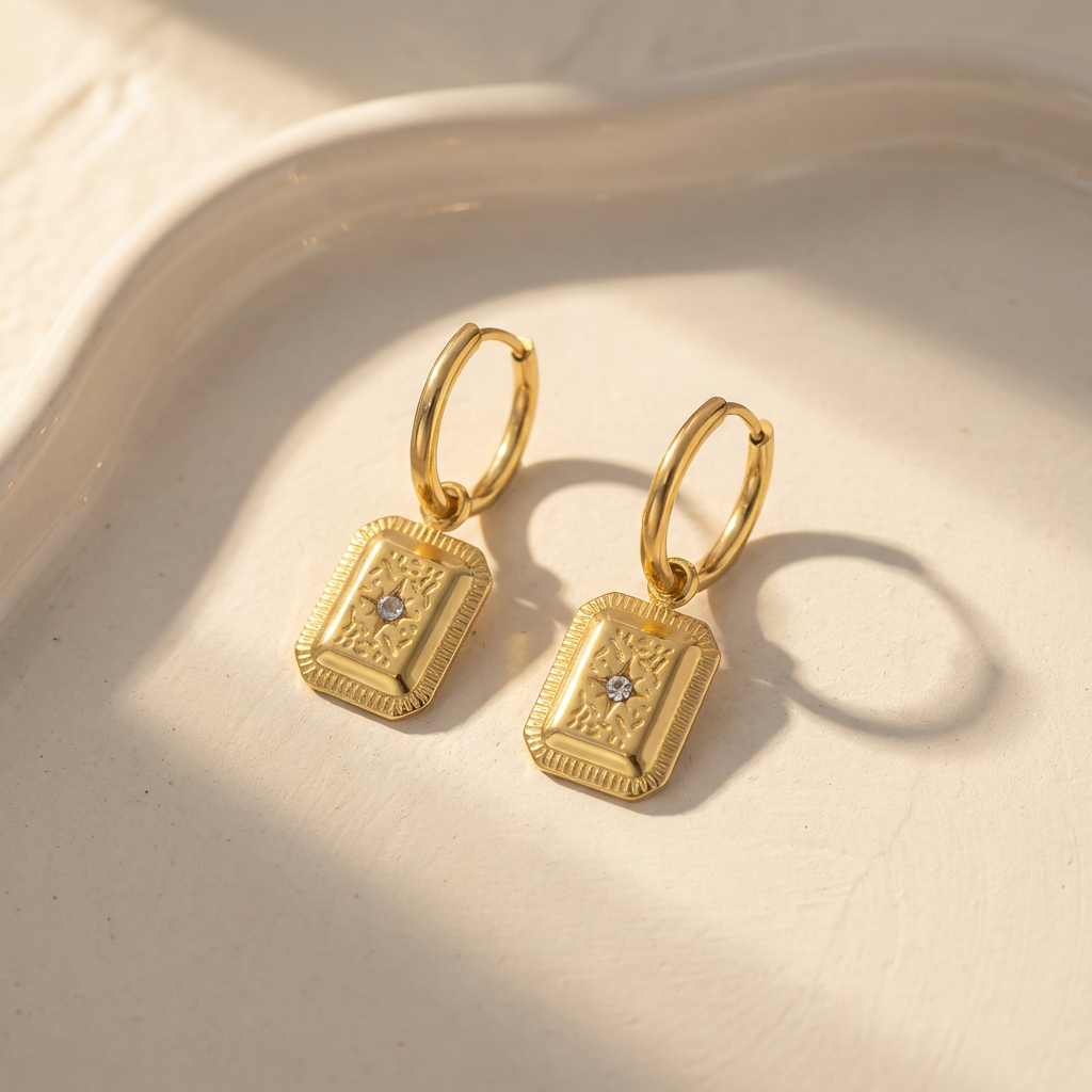  Gold tone stainless steel huggie hoop earrings with stamped rectangular charm and crystal detail on warm ivory ceramic backdrop in soft golden hour light