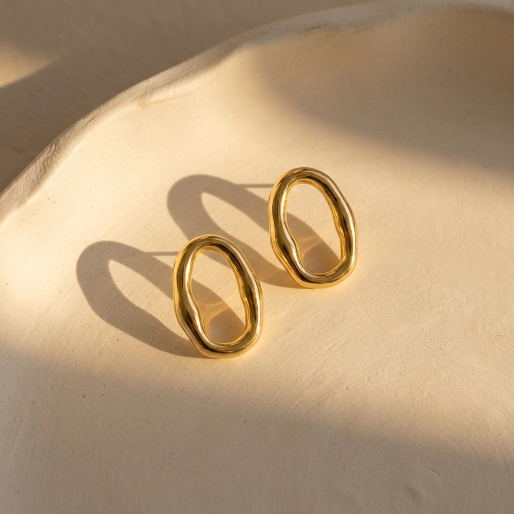 Close-up of gold organic oval hoop earrings with glossy shine on ivory plaster surface