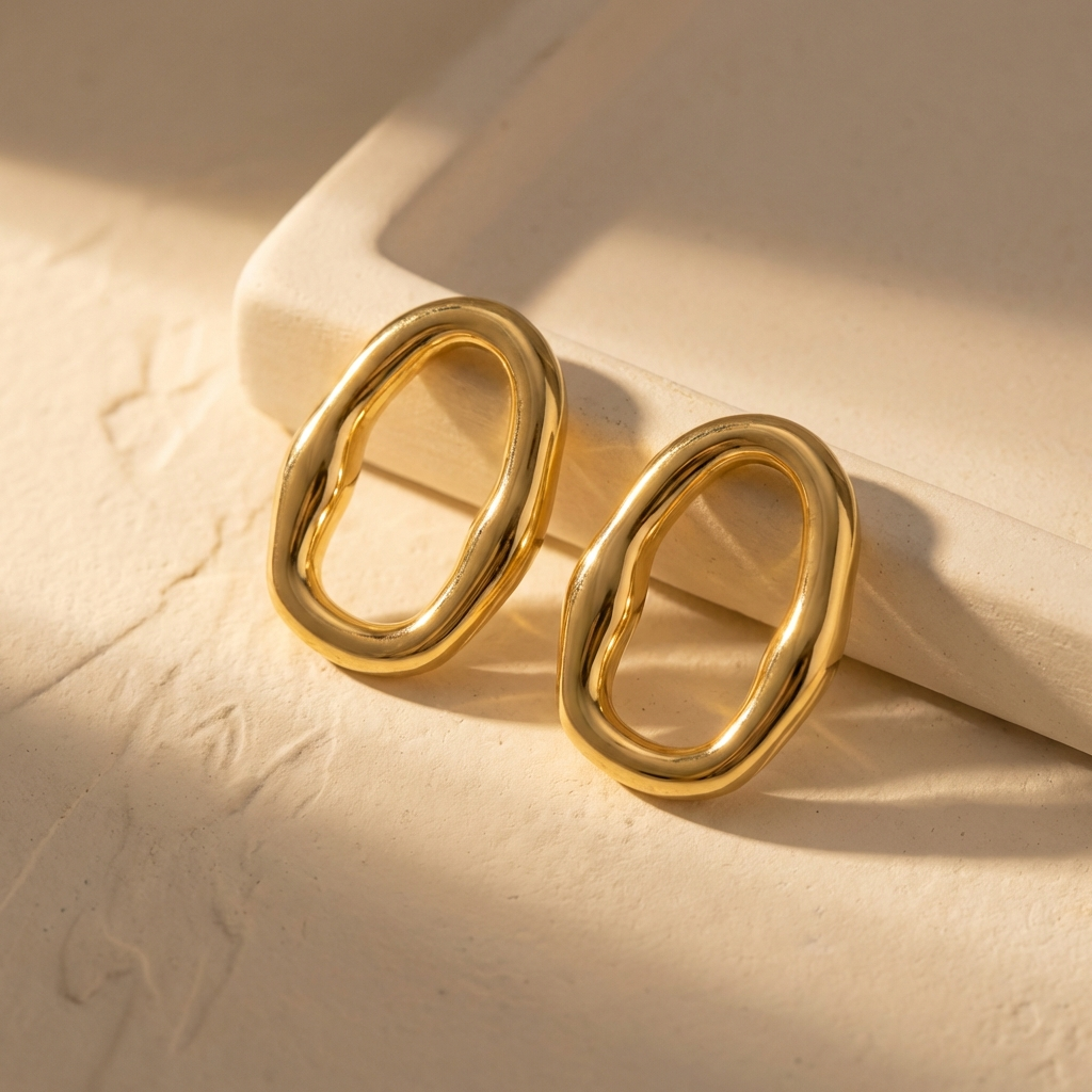 Gold organic oval hoop earrings on warm ivory ceramic backdrop in soft golden hour light